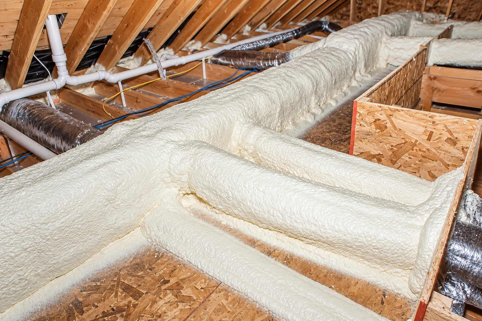 Thick white spray foam seals attic framing and ducts around wooden partitions and pipes.