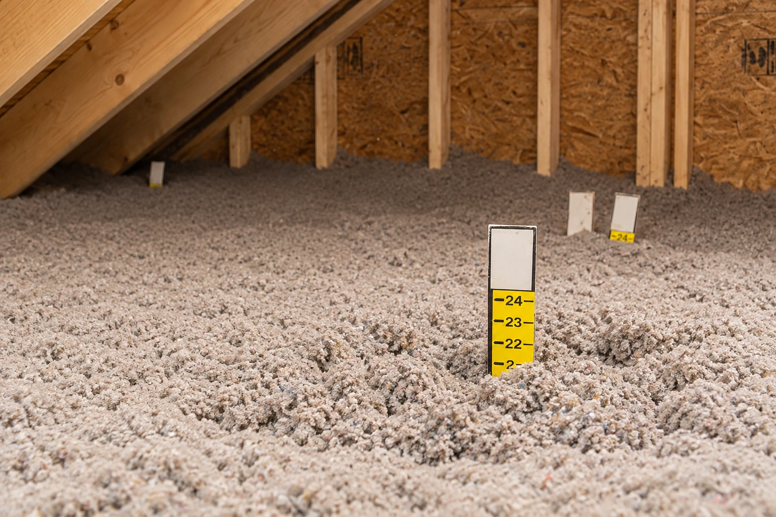Loose-fill insulation covers an attic floor, with a yellow depth marker protruding from the material.