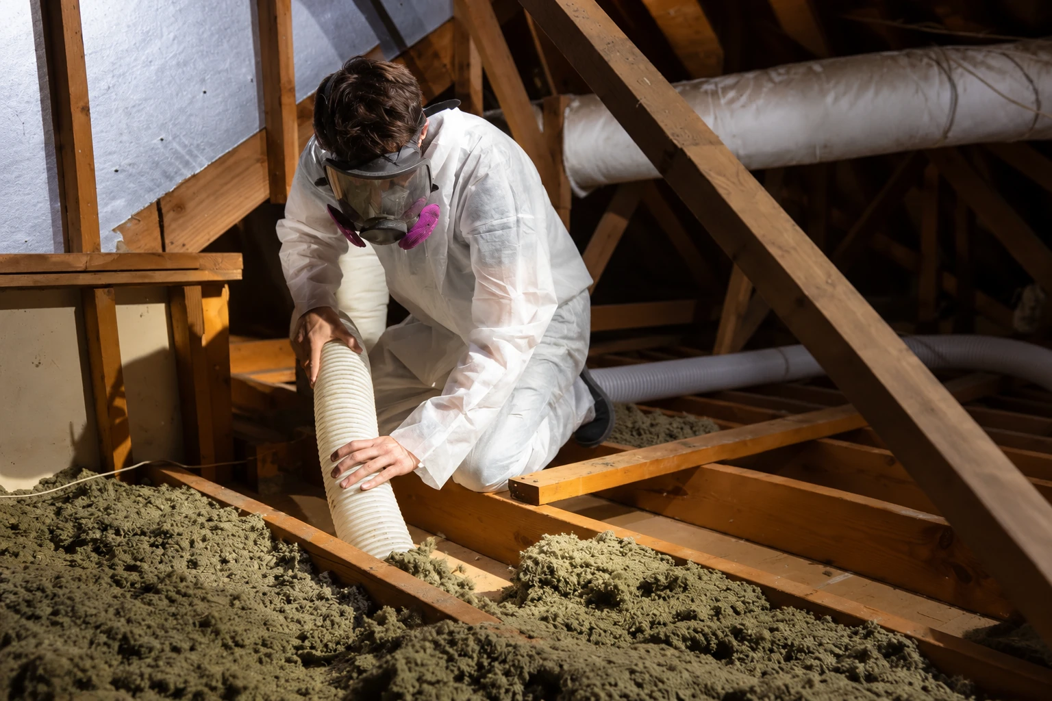 A worker in protective gear uses a wide hose to remove loose-fill insulation from an attic.