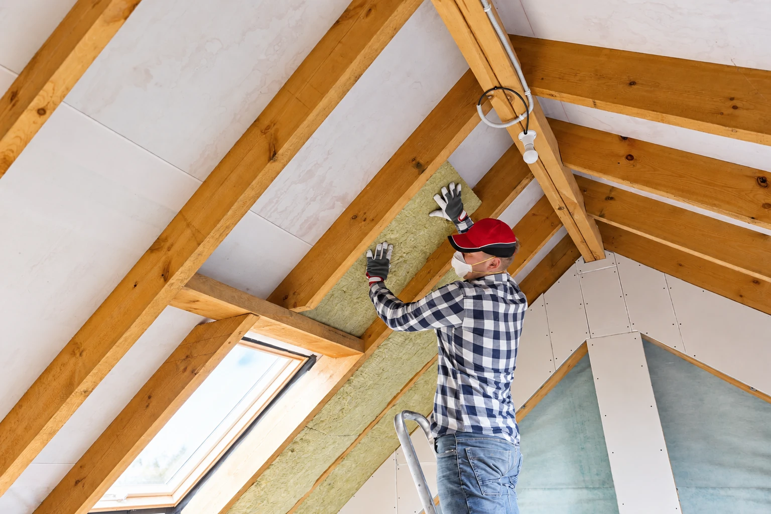 A worker in gloves and a mask fits a pale insulation batt between exposed wooden rafters.